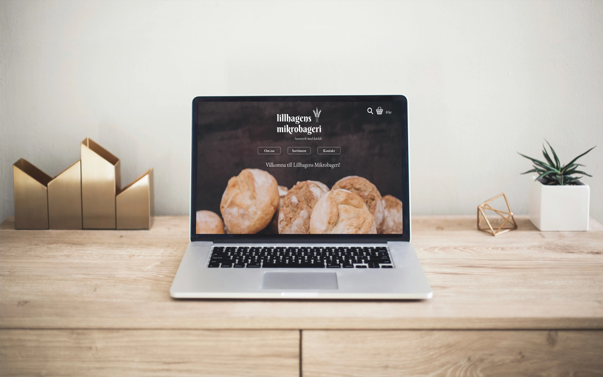 A laptop on a wooden desk showing the new Lillhagens Mikrobageri start page: logo, menu and “Välkomna till Lillhagens Mikrobageri!” over a photo of fresh bread.